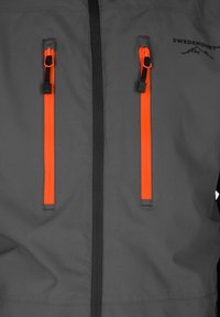 Grijze jas met oranje ritsaccenten en zwarte hardware. Glad materiaal met een matte afwerking, met een minimalistisch ontwerp.