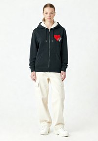 Zwarte hoodie met een schapenvachtvoering, voorzien van een rode hartafbeelding en veiligheidsklemmen. Gecombineerd met off-white cargo pants en witte sneakers.