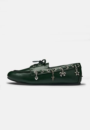 Mocassin en cuir vert foncé avec œillets argentés et quatre breloques pendantes : cœur, croix, nœud et étoile sur le côté.