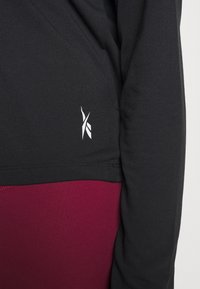 Black long-sleeve shirt with a fitted design, featuring a white logo on the bottom hem, paired with burgundy leggings. Smooth texture.