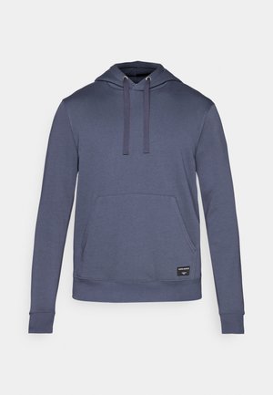 Tumši zila kapucīna jaka, kas izgatavota no mīksta auduma, ar priekšējo kabatu, regulējamiem auklām un nelielu zīmola etiķeti apakšmalā.