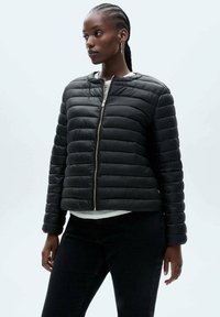 Zwarte pufferjack met horizontale stiksels, ritsluiting, ribgebreide manchetten en een cropped ontwerp. Gladde textuur en minimalistische stijl.