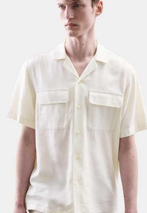 Ung mand iført en løs, kortærmet, cremefarvet skjorte med to brystlommer, der kigger let til siden.
