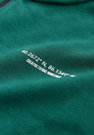 Material verde-albăstrui cu text alb: „40.2672° N. 86.1349° V” și „CREATIVE FUTURE INDUSTRIES” imprimat pe suprafață. Textură netedă, design casual.