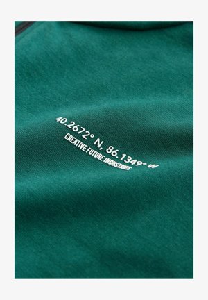 Material verde-albăstrui cu text alb: „40.2672° N. 86.1349° V” și „CREATIVE FUTURE INDUSTRIES” imprimat pe suprafață. Textură netedă, design casual.