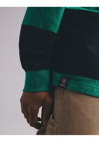 Zielony i granatowy sweter w paski z miękkiego materiału, z prążkowanym mankietem oraz metką z logo i napisem "1993" na dolnym brzegu. Ręka spoczywa na boku.