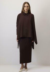 Blusa oversized marrone con colletto a punta, bottoni sul davanti e design fluido, abbinata a una gonna alta coordinata.