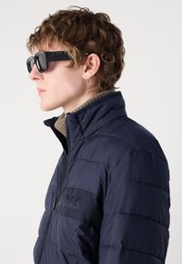 Jūrinis pūkinis švarkas turi tekstūruotą paviršių, užtrauktuku užsegamą priekį ir iškilusį logotipą ant krūtinės; derinamas su dideliais juodais saulės akiniais.