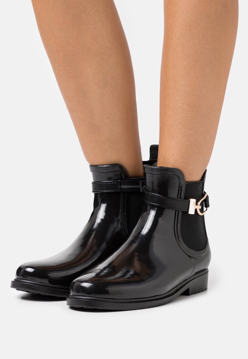 Bottes noires en vinyle à la cheville avec une finition brillante, des panneaux latéraux élastiques et une bride décorative avec un accent métallique. Semelle plate et bout arrondi.