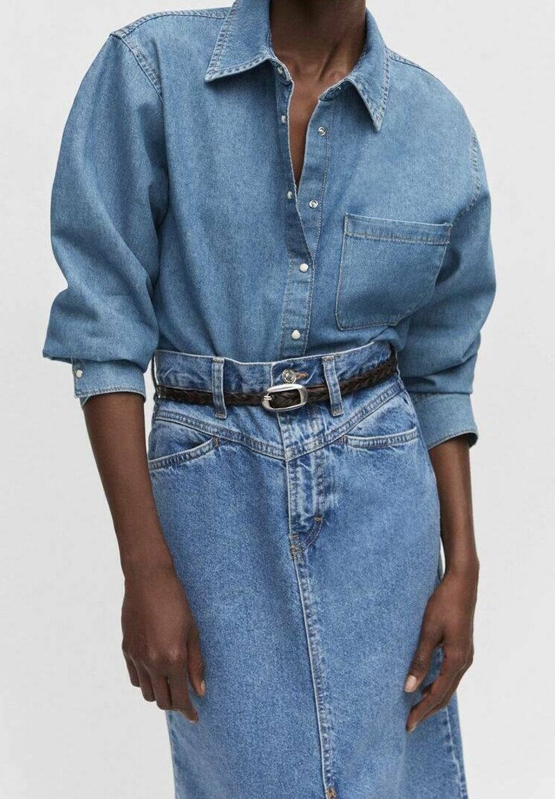 Personne à la peau foncée portant une chemise en jean bleue boutonnée rentrée dans une jupe en jean taille haute avec une ceinture tressée marron foncé.