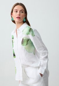 Camicia bianca in lino con grandi stampe di foglie verdi su un lato. Maniche lunghe con polsini a bottone, vestibilità rilassata, infilata in pantaloni bianchi.