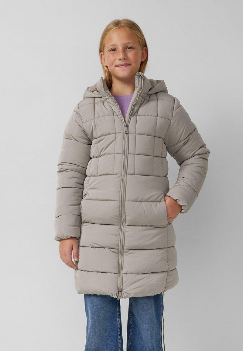 Djevojka nosi svijetlo sivi zimski kaput s kapuljačom, ljubičastu majicu ispod i plave traperice, stoji naslonjena na svijetlu pozadinu.