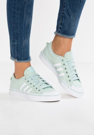 Voeten met lichtgroene sneakers met witte strepen en witte zolen, gecombineerd met blauwe jeans met rafelige zoom op een witte vloer.