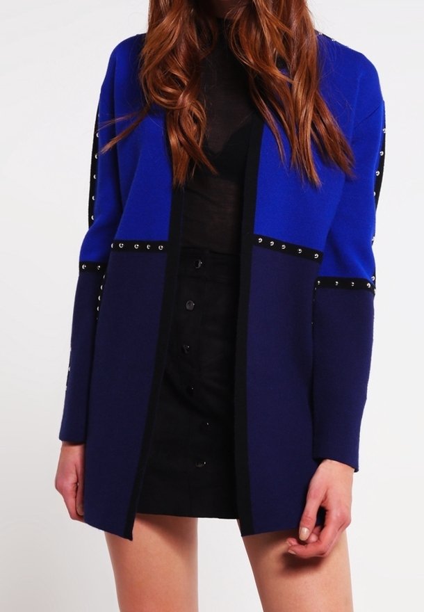 Blauer Cardigan mit schwarzen Akzenten, strukturiertem Design, Nietendetails an den Ärmeln und geradem Schnitt, kombiniert mit einem schwarzen Rock.