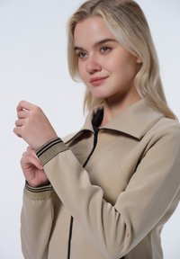 Beige sako s límcem, se černým zipem a texturovanými manžetami se dvěma černými pruhy. Hladká látka, střih na míru.