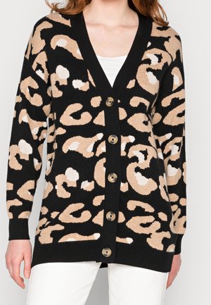 Femme portant un cardigan noir avec un imprimé léopard beige et blanc, boutonné sur le devant, par-dessus un haut blanc et un pantalon blanc.