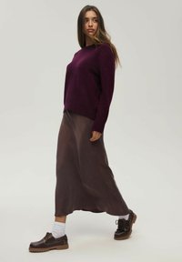 Fioletowy dzianinowy sweter, długa brązowa satynowa spódnica oraz ciemne sznurowane buty z grubą podeszwą. Modelka stoi bokiem, prezentując warstwowe tekstury.