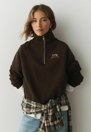 Kvinde iført en mørkebrune sweatshirt med halvlange lynlås og teksten "cosy season", lyseblå jeans og en ternet skjorte bundet om taljen.