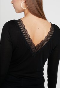 Černý top s dlouhými rukávy, který má na zadní straně V-špici s krajkovým lemem a složitými vzory, vyrobený z měkkého materiálu. Jednoduchý a přiléhavý design.