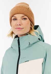 Mintgroene jas met witte paneel, voorzien van een hoge kraag en rits. Bruine gebreide beanie met textuurpatroon en logo-label.