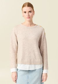Maglione leggero beige in maglia con orlo a coste, colletto a giro e bottoni decorativi sulla spalla. Indossato sopra una camicia bianca con colletto.