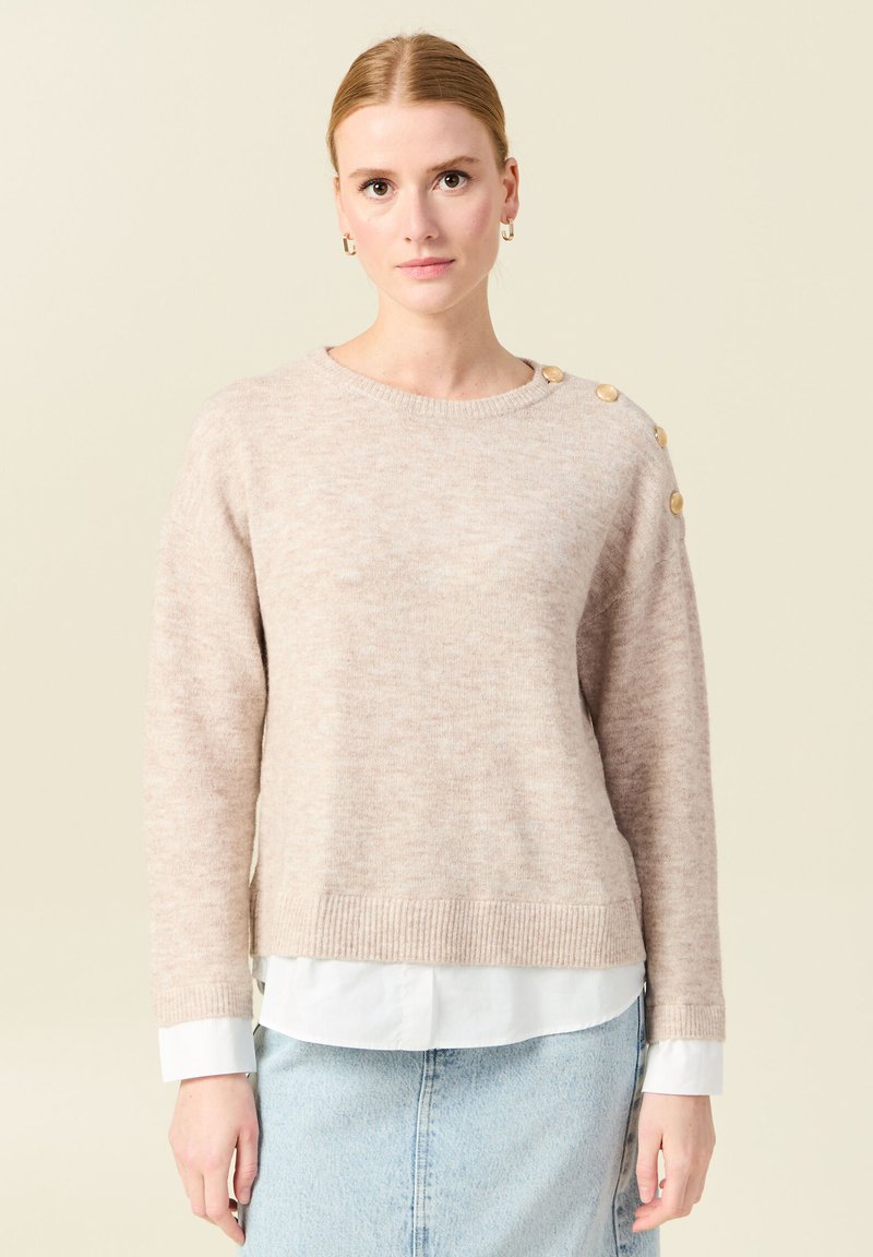 Maglione leggero beige in maglia con orlo a coste, colletto a giro e bottoni decorativi sulla spalla. Indossato sopra una camicia bianca con colletto.