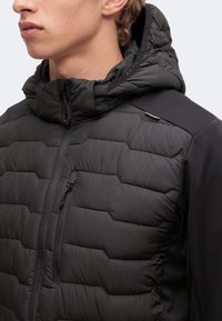 Jeune personne portant une veste matelassée noire avec capuche, col montant et poche zippée visible sur la poitrine, faisant face légèrement vers la gauche.
