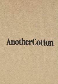 Beige katoenen stof met een textuurafwerking heeft de tekst "AnotherCotton" in vette, zwarte, serif-lettertype gecentreerd op het oppervlak.