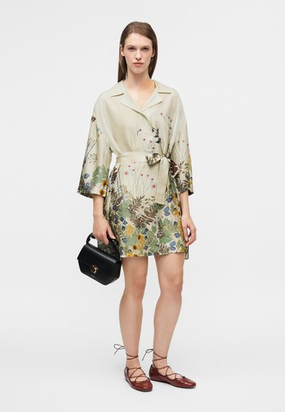 Robe courte fleurie en tissu léger, avec des manches larges, une ceinture et un mélange de couleurs vert, jaune et rose. Associée à un sac noir et des ballerines rouges.