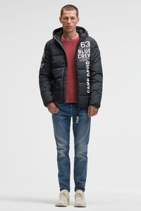 Man met een zwarte gewatteerde capuchonjas met witte tekst, rode trui, blauwe spijkerbroek en witte sneakers, staand tegen een effen achtergrond.