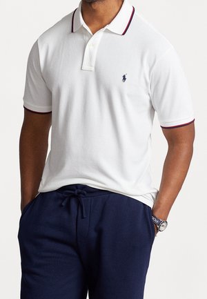 Hvid polo skjorte med en knapkrave, røde og marineblå accenter på ærmerne og et lille broderet logo på brystet. En stof af bomuld.