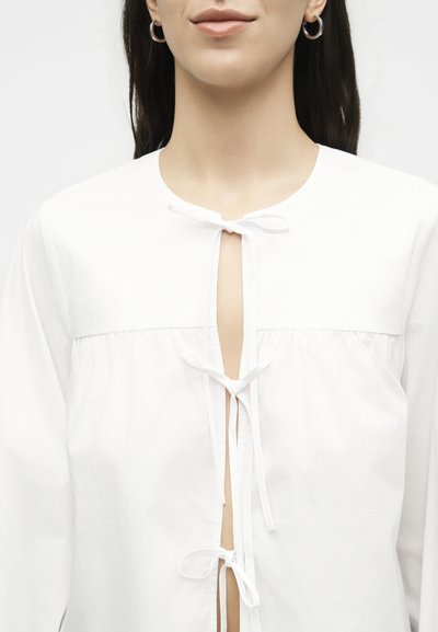 Γυναίκα που φοράει ένα λευκό μπλουζάκι με τρία δεμένα φιόγκους μπροστά και μικρά σκουλαρίκια κρίκους, σε απλό ανοιχτό φόντο.