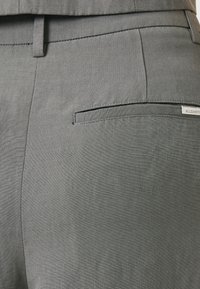 Gros plan sur un pantalon de costume gris montrant une poche passepoilée arrière avec une petite étiquette de marque cousue près du bord droit.