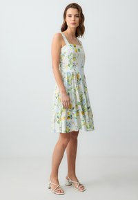 Bloemenpatroon zomerjurk met een vierkante halslijn, lichte stof, gelaagde rok en groene en gele accenten; gecombineerd met witte sandalen met banden.