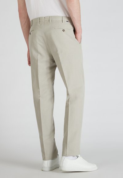 Pantalon beige clair avec une texture lisse, coupe ajustée, deux poches arrières avec fermetures à bouton, porté avec des baskets blanches.
