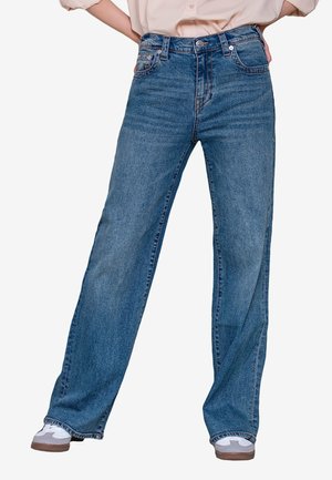 Persona che indossa jeans blu a gamba larga e vita alta e scarpe da ginnastica bianche, in piedi con le mani sui fianchi davanti a uno sfondo semplice.