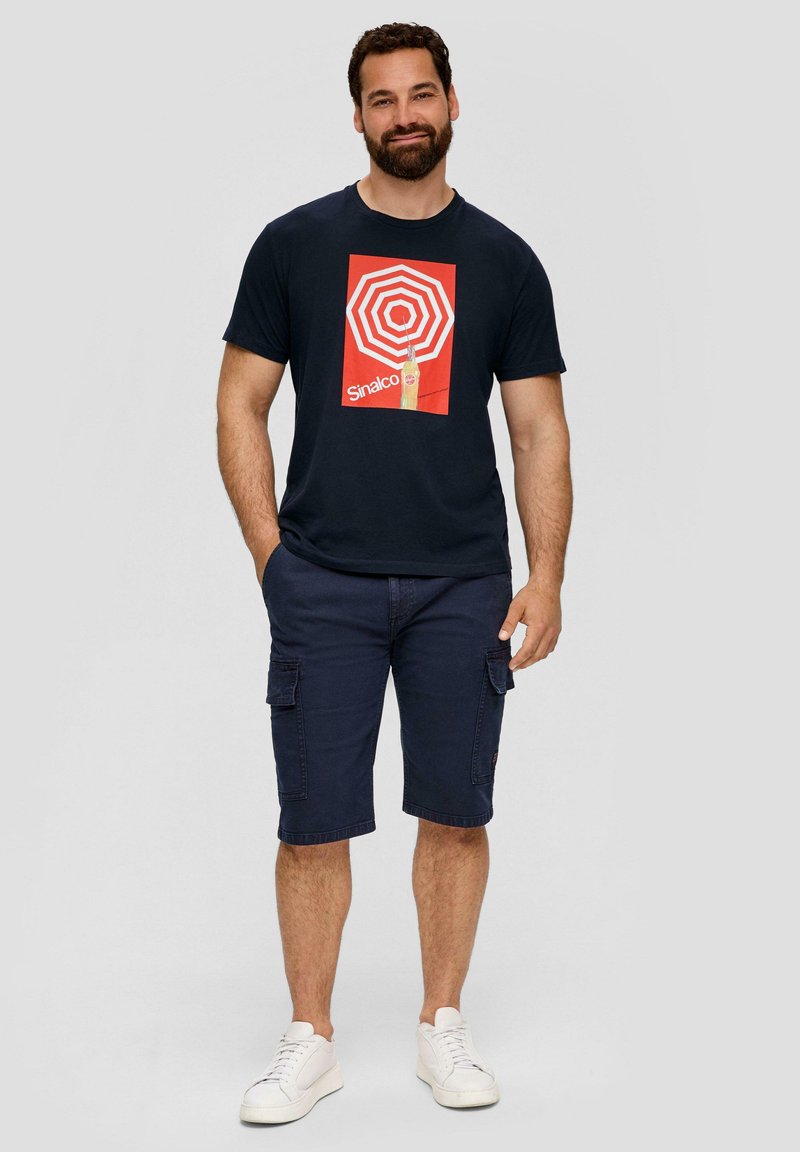 Navyblauwe katoenen t-shirt met een grafische print van rode en witte concentrische cirkels. Gecombineerd met donkere cargoshorts en witte sneakers.