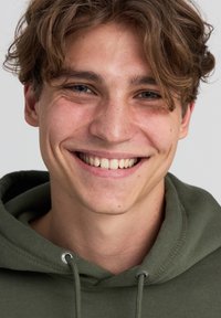Giovane uomo con capelli castani mossi che indossa un felpa verde. Ha lentiggini, un sorriso luminoso e una pelle liscia. Sfondo semplice.