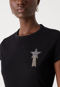Black cotton t-shirt featuring a star patch embellished with rhinestones and fringe details, positioned on the left shoulder.