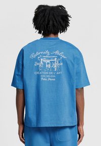 Sininen puuvillapaita, jossa on rento istuvuus. Paidassa on valkoista tekstiä ja graafinen kuva takana, johon kuuluu "Reternity Atelier" ja yhteystiedot.