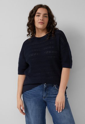 Vrouw met krullend haar die een marineblauwe gebreide trui met korte mouwen en een blauwe spijkerbroek draagt, staand tegen een effen achtergrond.