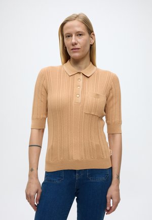 Blonde vrouw die een beige gebreide polo met korte mouwen en een borstzak draagt, gecombineerd met een blauwe spijkerbroek, staand tegen een effen witte achtergrond.