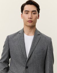 Γκρι σακάκι με ρίγες από υφή υφάσματος, με κλείσιμο με μία κουμπί, κανονικούς γιακάδες και τσέπη στο στήθος. Φορεμένο πάνω από λευκό πουκάμισο.