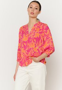 Bluse mit floralem Druck in leuchtendem Pink und Orange, mit Dreiviertelärmeln, einem V-Ausschnitt und aus leichtem Material. Kombiniert mit weißen Hosen.