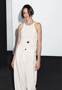 Witte, mouwloze tanktop van een zachte stof, gecombineerd met losse, hoge witte broeken met een zwarte knop. Lange zilveren ketting met een ronde hanger.