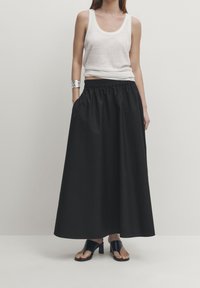 Gonna maxi nera con elastico in vita e tasche laterali, abbinata a un top bianco senza maniche. Il modello indossa sandali neri con tacco a blocco.