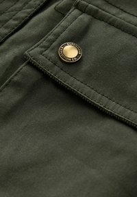 Närbild av en mörkgrön tygficka med sömmar och en mässingsknapp märkt "Superdry Expedition Co."