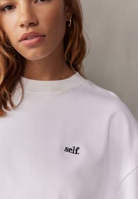 Vit sweatshirt i bomull med avslappnad passform. Har en liten broderad "self." logotyp i svart. Slät textur och rundad halsdesign.