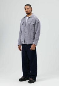 Grijze, gestructureerde fleecejas met knoopsluiting, twee borstzakken en een kraag. Draagt over een wit shirt met donkere spijkerbroek.