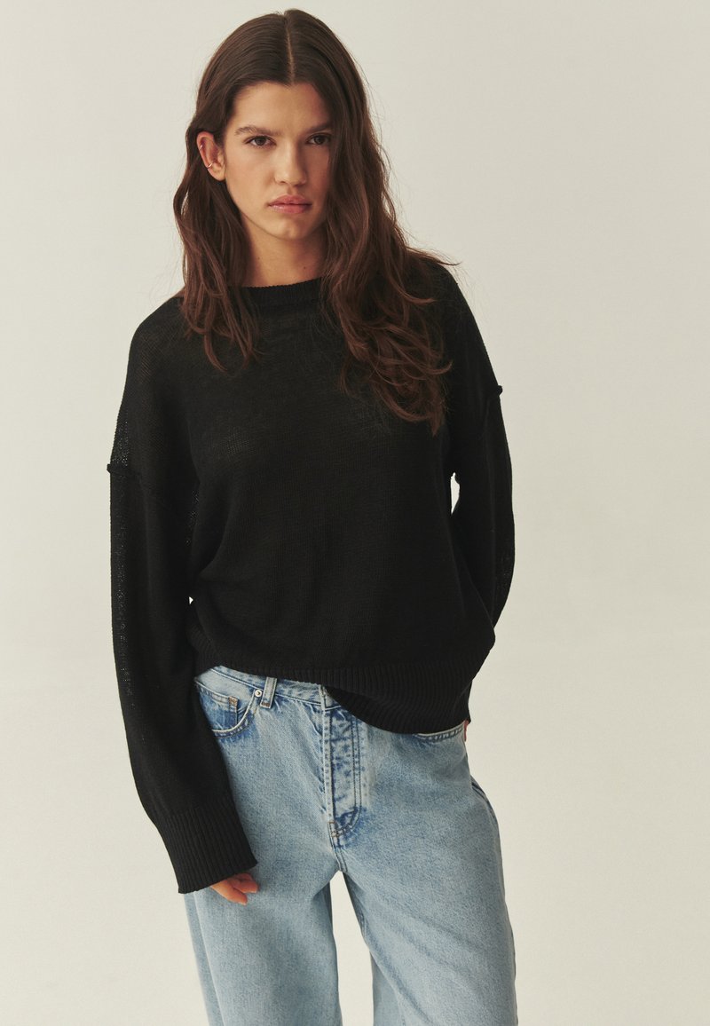 Jeune femme aux longs cheveux bruns portant un pull noir et un jean bleu clair, debout devant un fond beige uni.